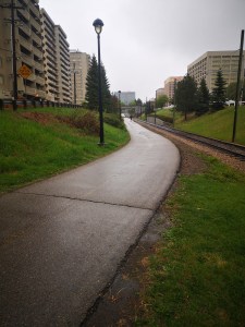Multi-use paved trail running through a green space. Apartment and office buildings visible on either side of the green space