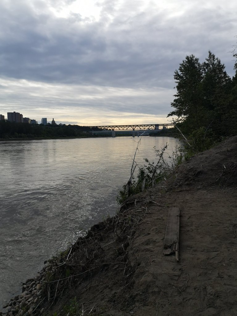 A muddy bank leads to a dark river. Two bridges are in the distance 