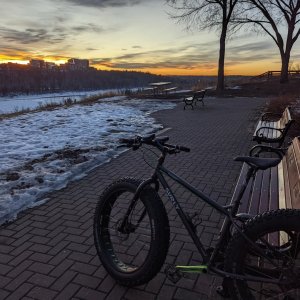 A grey fat bike with green cranks leans against a park bench overlooking a snow covered river valley. A sunset fades in the background